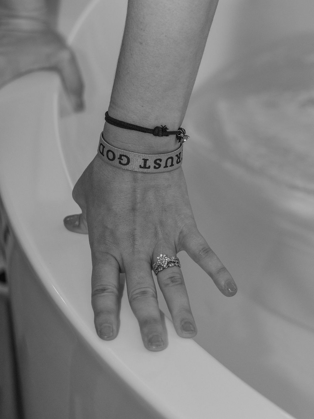 Black and white photo of a hand resting on a bathtub edge, wearing a 'Trust God' bracelet and a decorative ring.