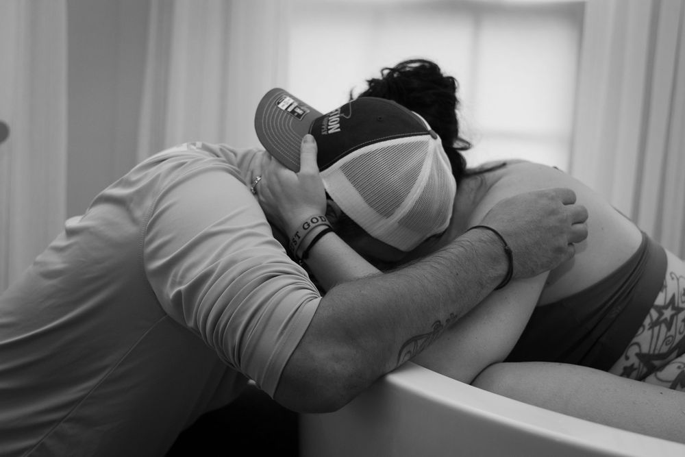 Black and white photo of a couple embracing during labor, woman wearing a medical mask leaning on a birthing tub.