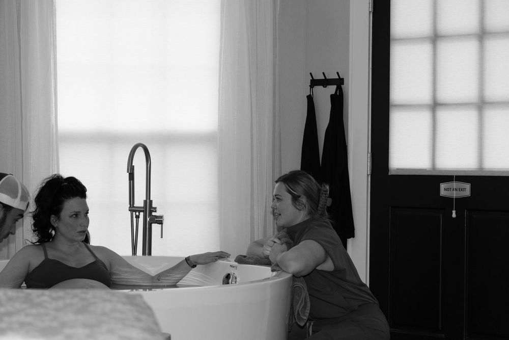Black and white photo of a woman relaxing in a freestanding bathtub assisted by two caregivers indoors.