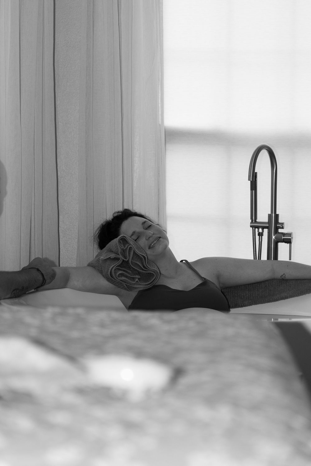 Black and white photo of a woman relaxing in a bathtub with a floral eye mask, near a window.