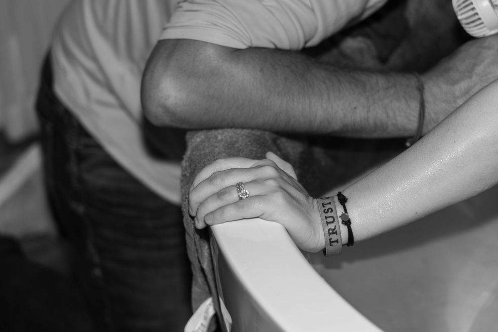 Close-up black and white photo of a couple holding hands over a white birthing tub during labor.
