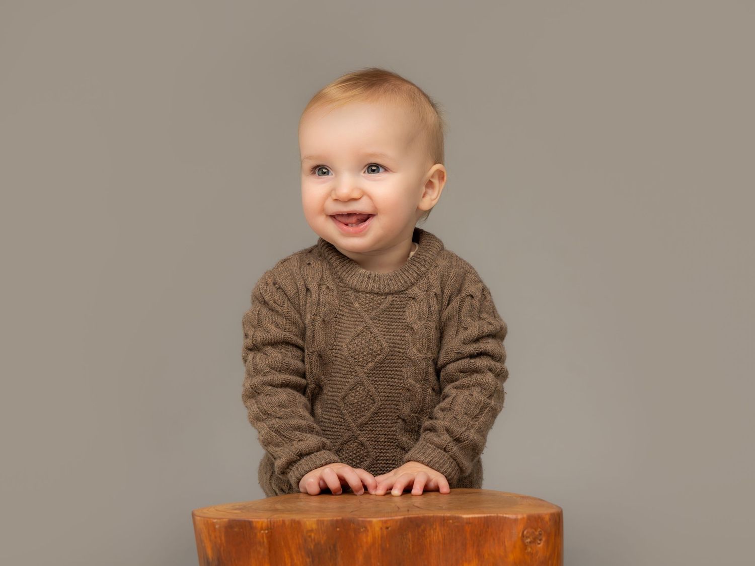 Smiling baby in brown cable-knit sweater leaning on wooden stool against gray background.