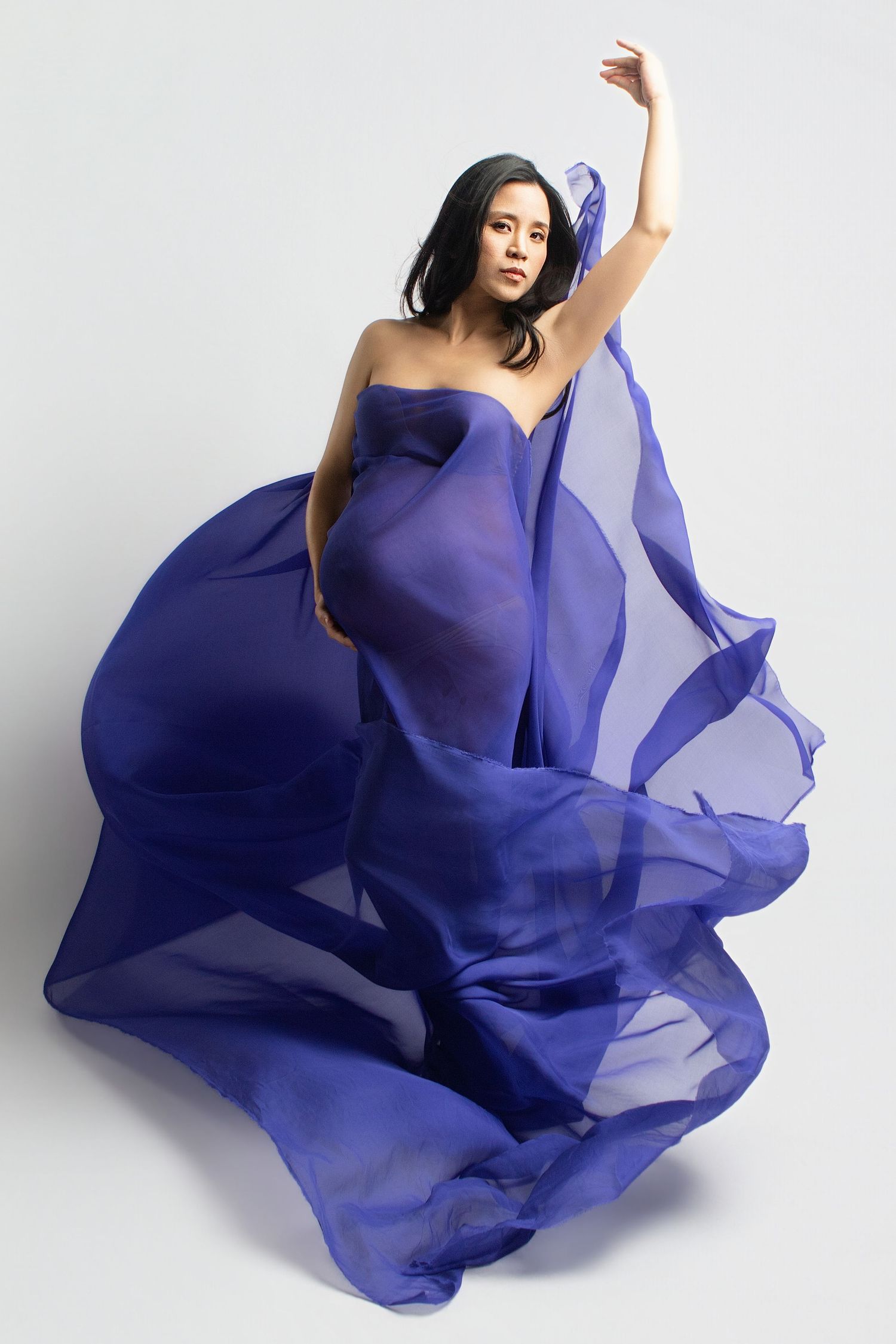 Pregnant woman in flowing blue chiffon fabric gown posing gracefully against white background.