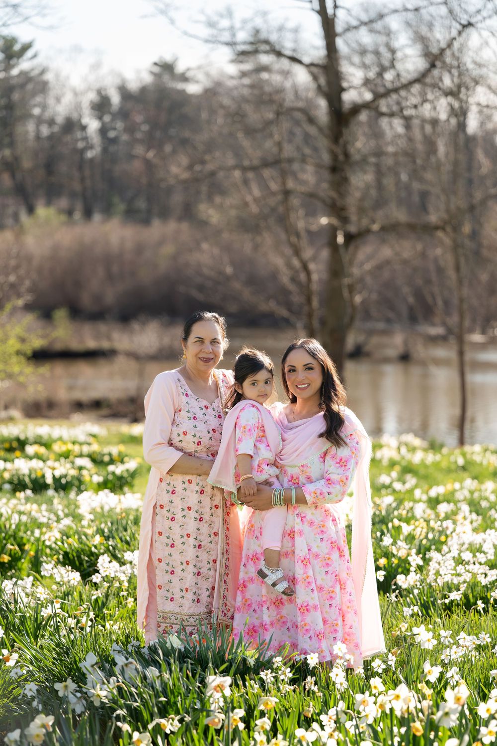 Three generations of women in coordinating pink floral outfits standing in daffodil meadow.