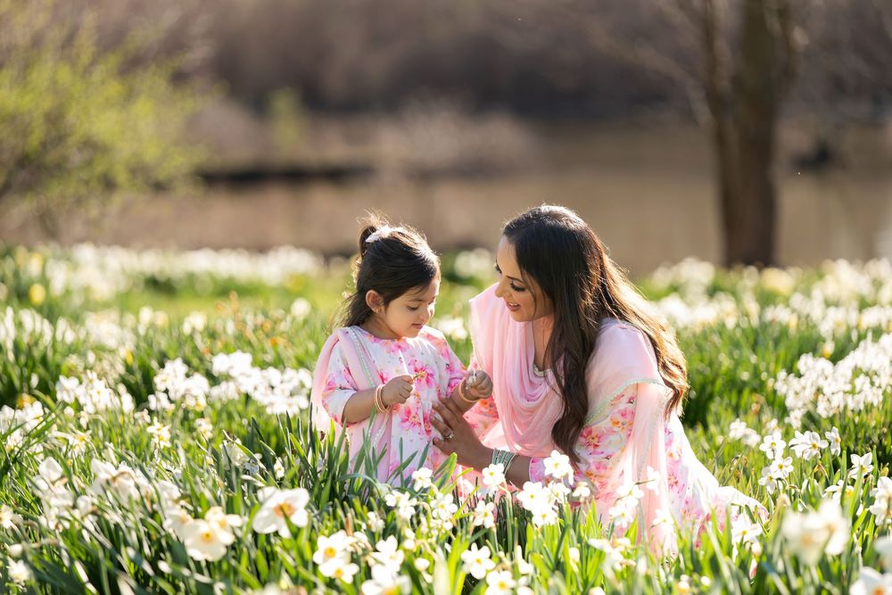 Mother in pink sari and daughter in floral dress exploring white daffodils in a sunny field.