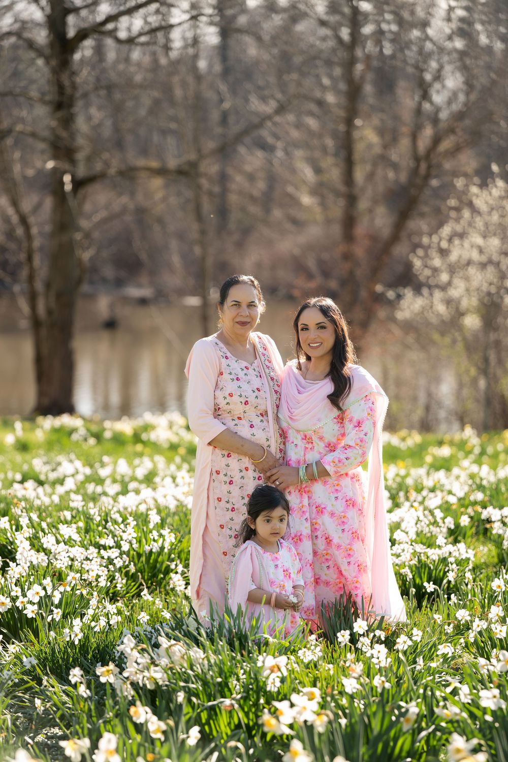 Three generations of Indian women in pink traditional outfits posing in daffodil field by pond.