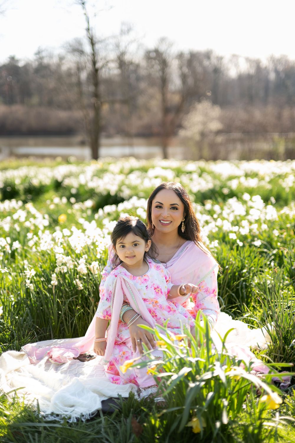 Mother and daughter in pink Indian outfits sitting together in lush daffodil field on sunny spring day.