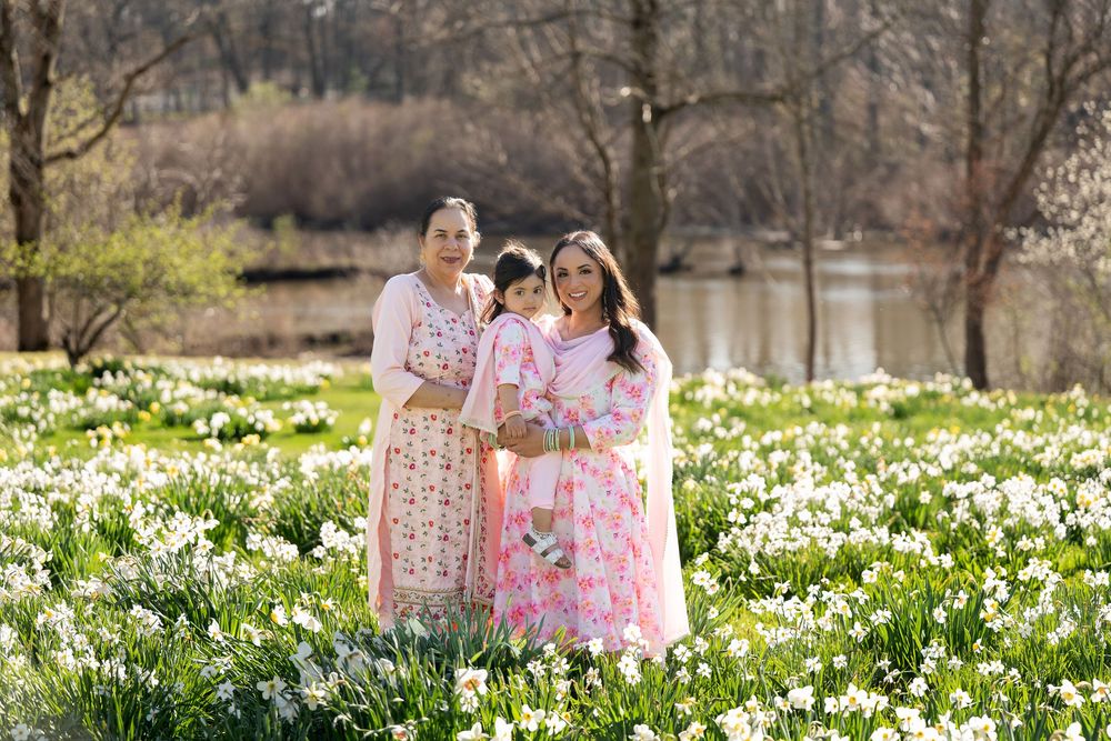 Three generations of women in pink floral attire standing among white daffodils near a pond.