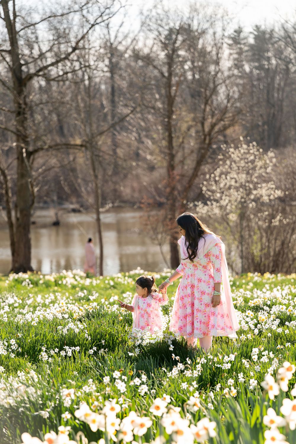 Mother and young daughter in matching pink dresses walking through a blooming daffodil field near a lake.