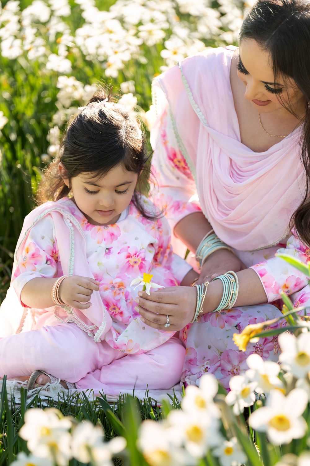 Young girl and mother in pink Indian outfits examining a flower while sitting in a daffodil field.