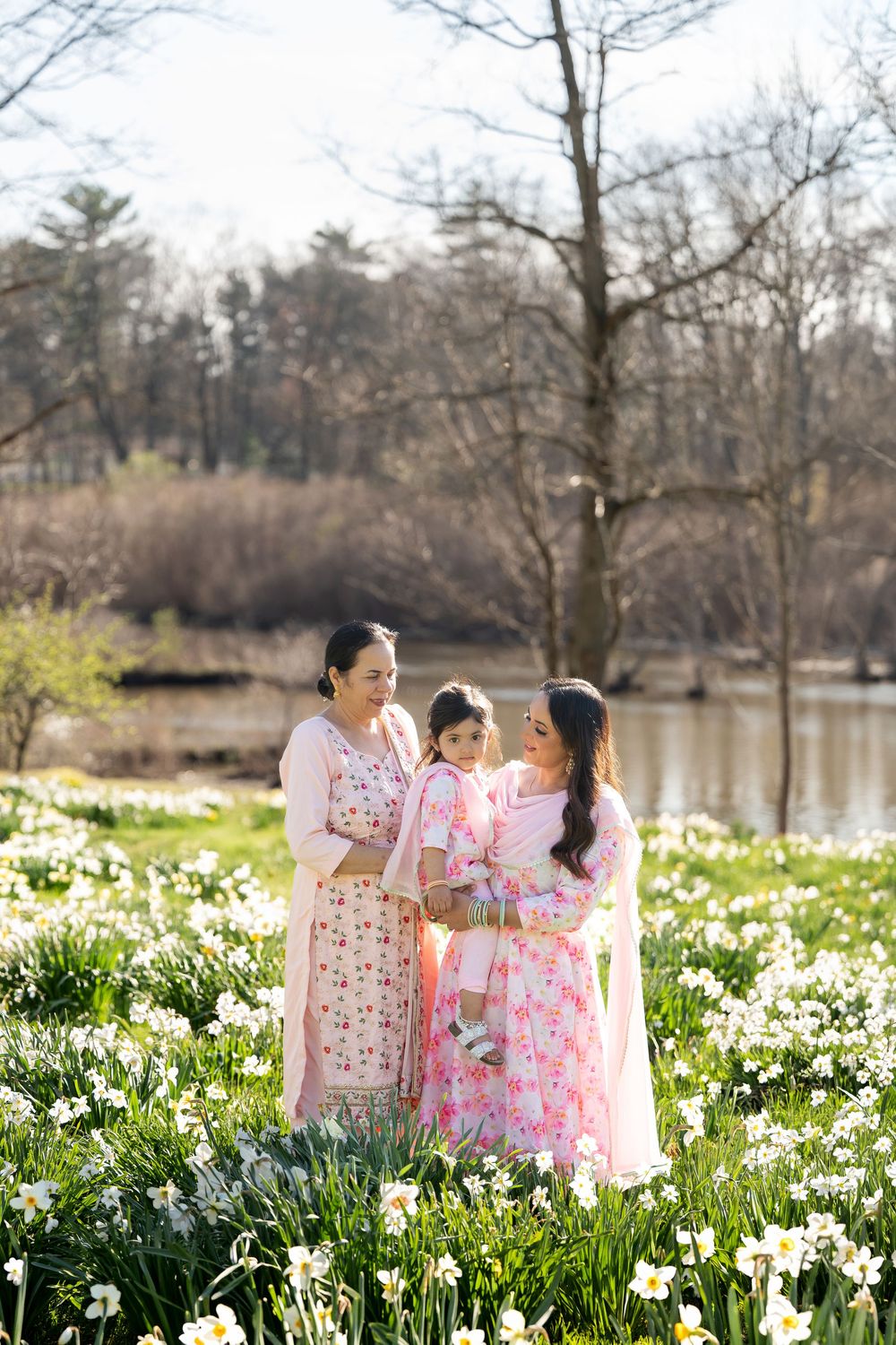 Three generations in pink floral outfits standing among white daffodils by a pond in spring.