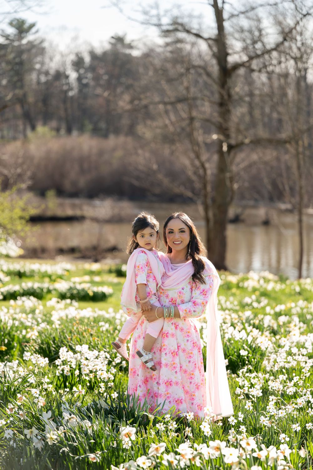Mother in pink floral sari holding young daughter in matching outfit in a daffodil field.