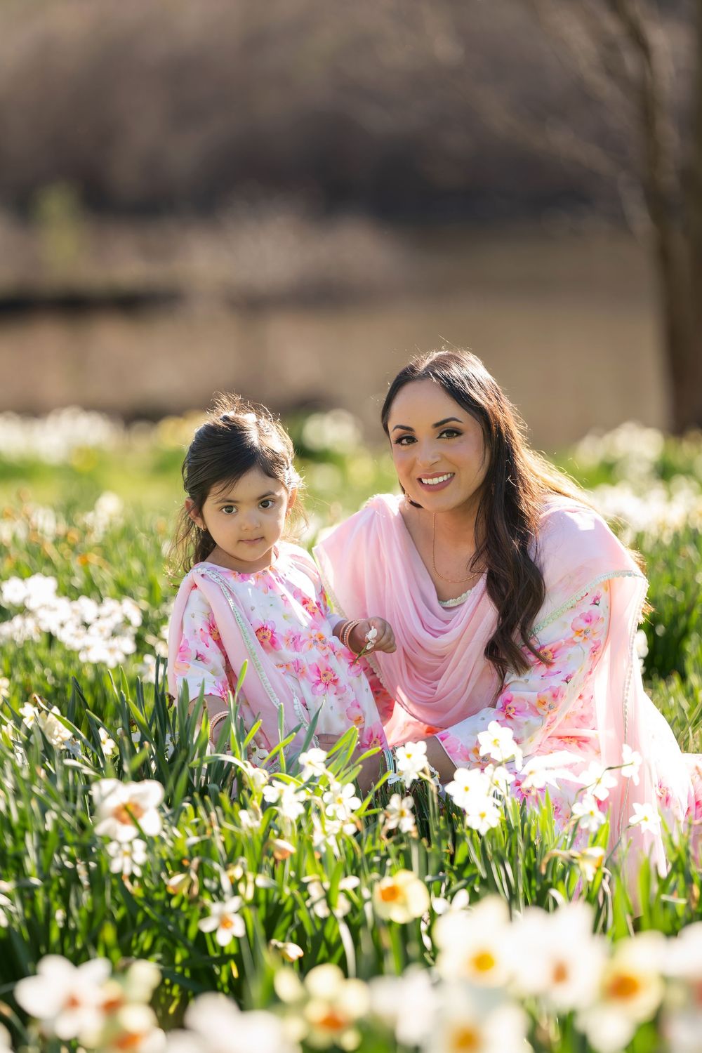 Smiling mother and young daughter in pink outfits posing among white spring daffodils outdoors.