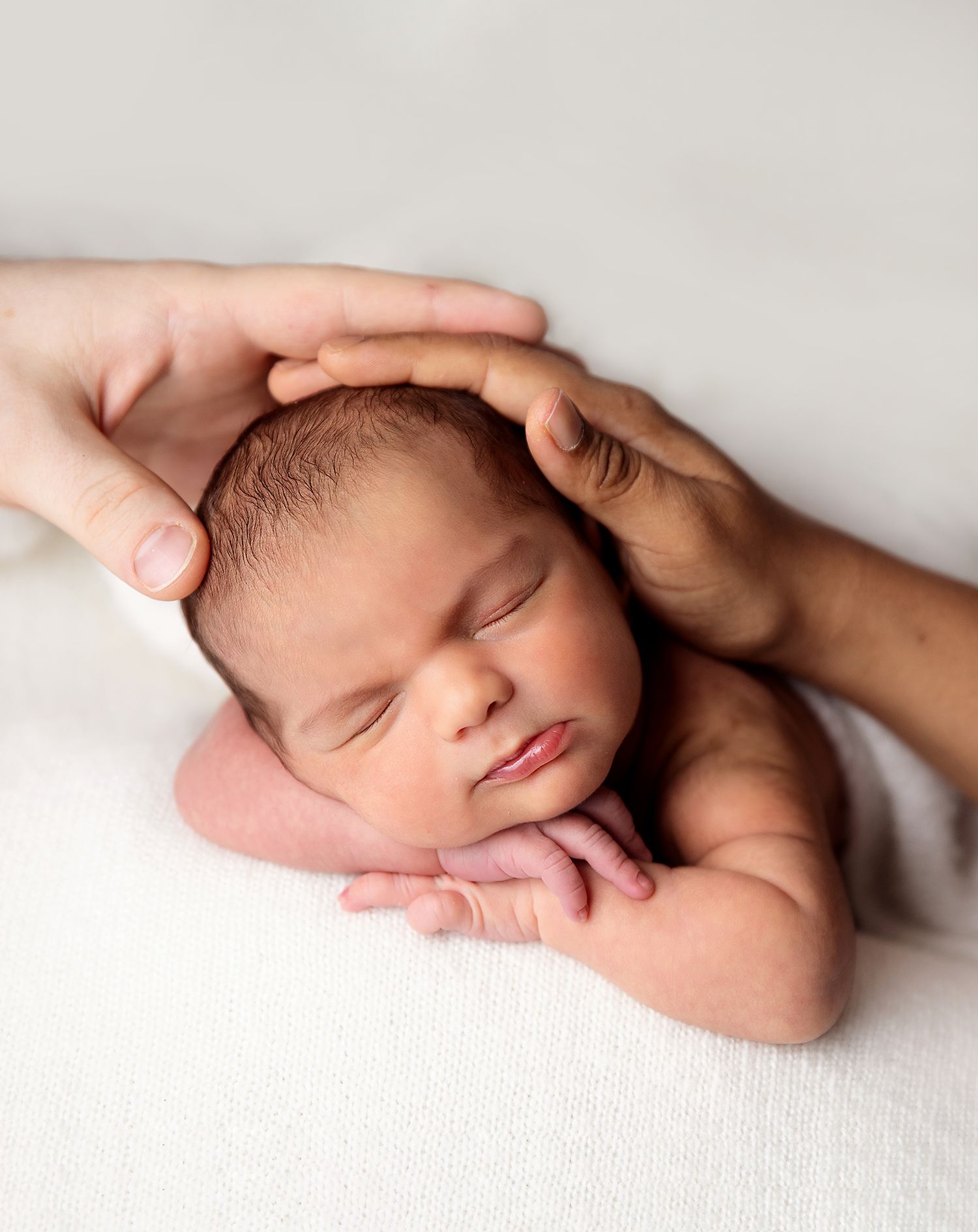 Newborn baby sleeping peacefully with parents hands cradling head on white blanket.