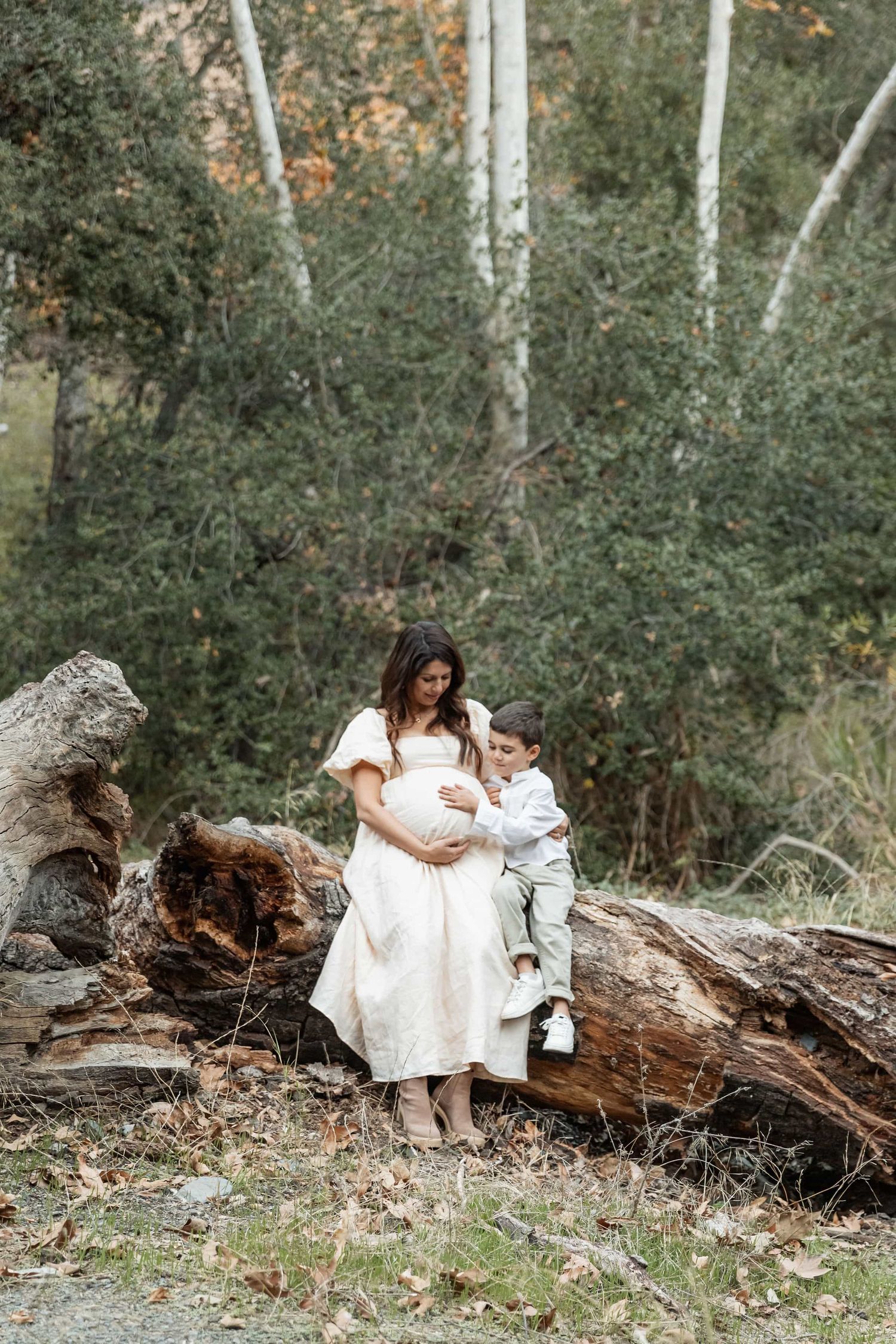 Pregnant woman in white dress with young son beside a fallen log in forest.