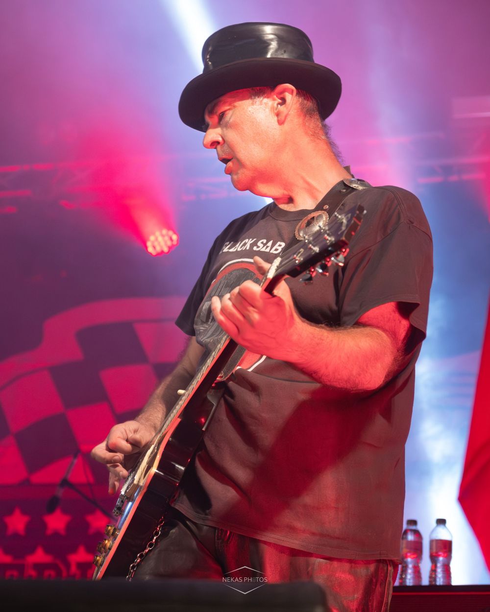 Guitarist in black top hat and Black Sabbath t-shirt performs on stage under red lights.