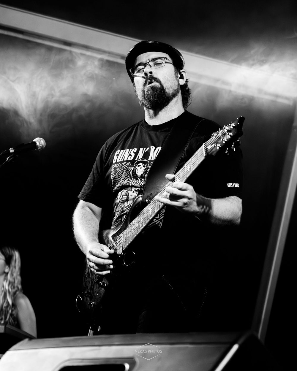 Black and white photo of a bearded male guitarist performing on stage wearing a Guns N Roses t-shirt and cap.