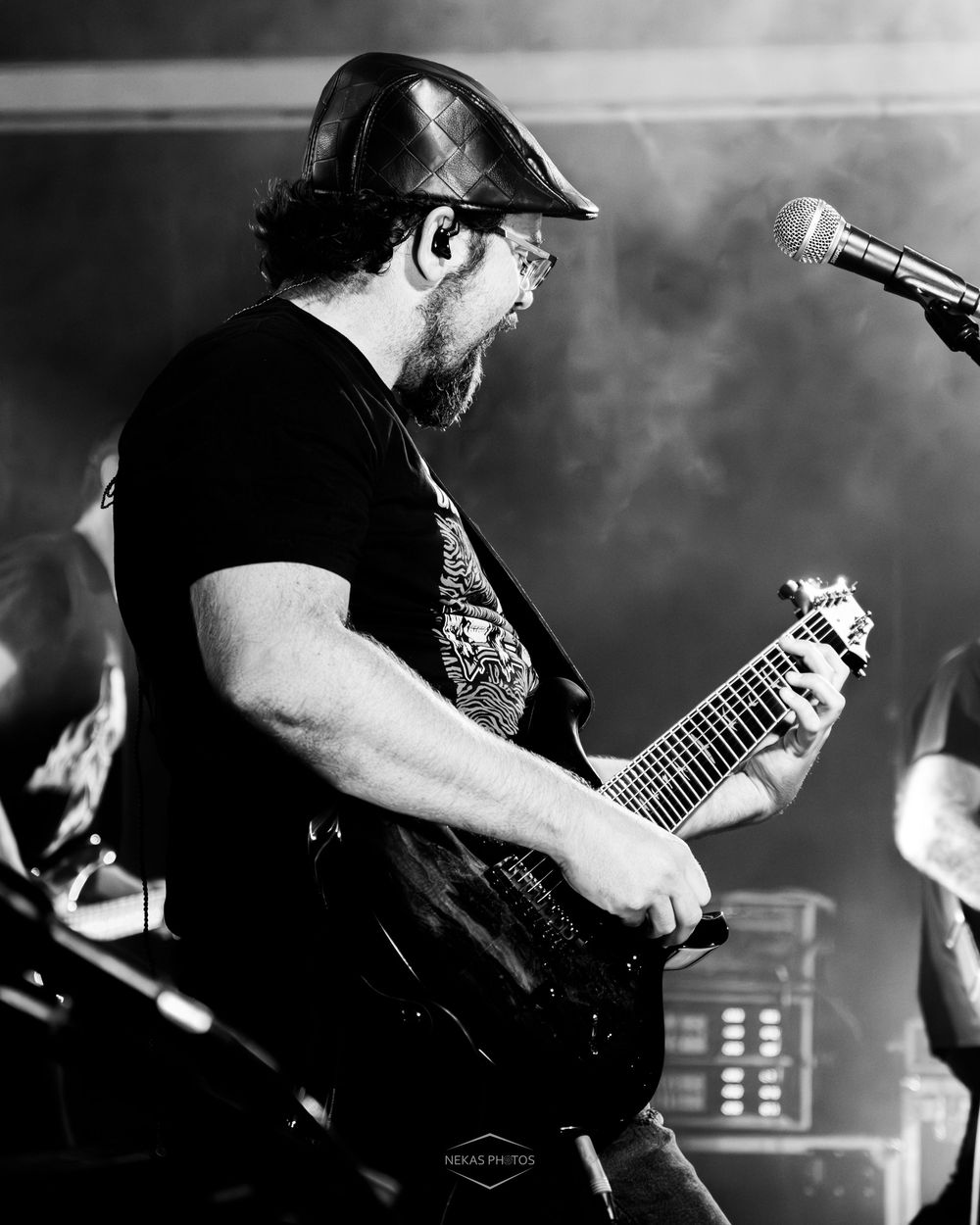 Black and white photo of a bearded guitarist performing on stage wearing a cap, playing electric guitar near a microphone.
