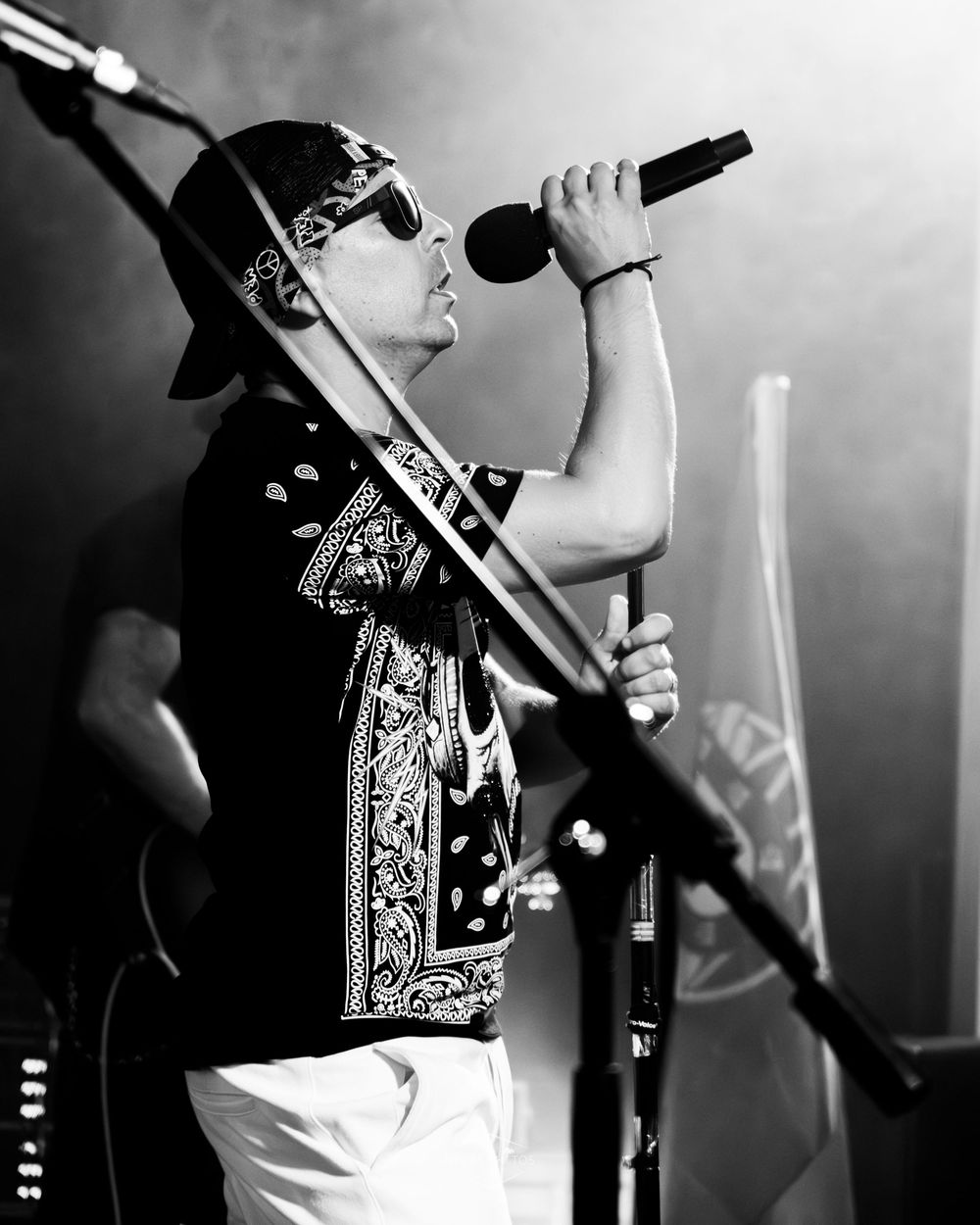 Black and white photo of a male singer performing on stage, holding a microphone, wearing a bandana and cap.