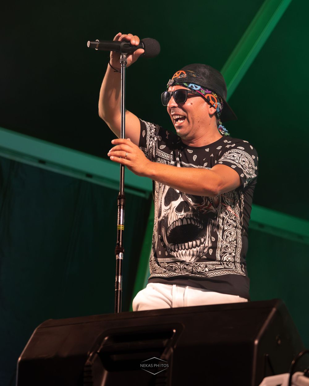 Male singer performing on stage wearing sunglasses, bandana headband, and skull print shirt while singing into microphone.