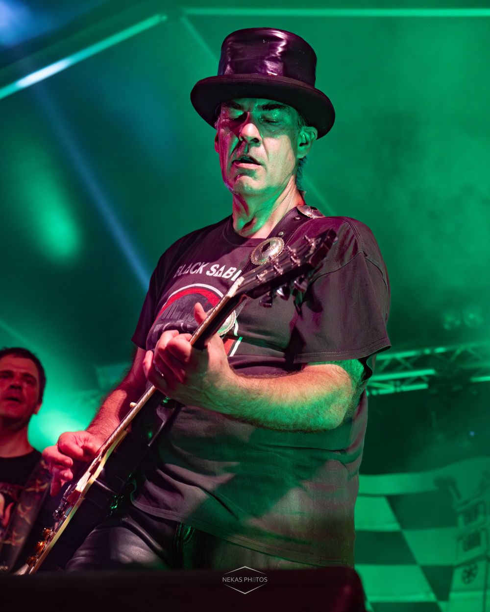 Guitarist in a top hat and Black Sabbath shirt performs on stage under green lights at a live concert.