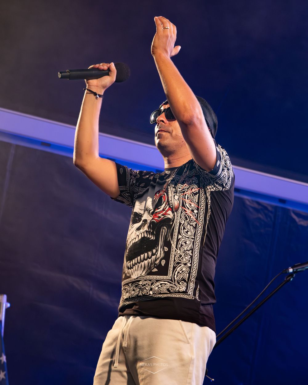 Male performer singing on stage, holding microphone up with one hand raised, wearing skull print t-shirt and cap.