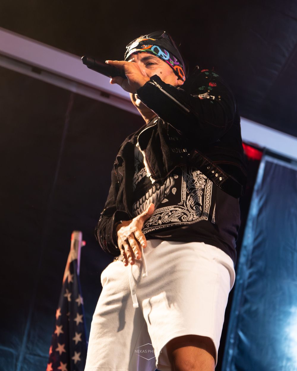 A rapper performs on stage holding a microphone, wearing white shorts, black jacket, and a cap with an American flag in the background.