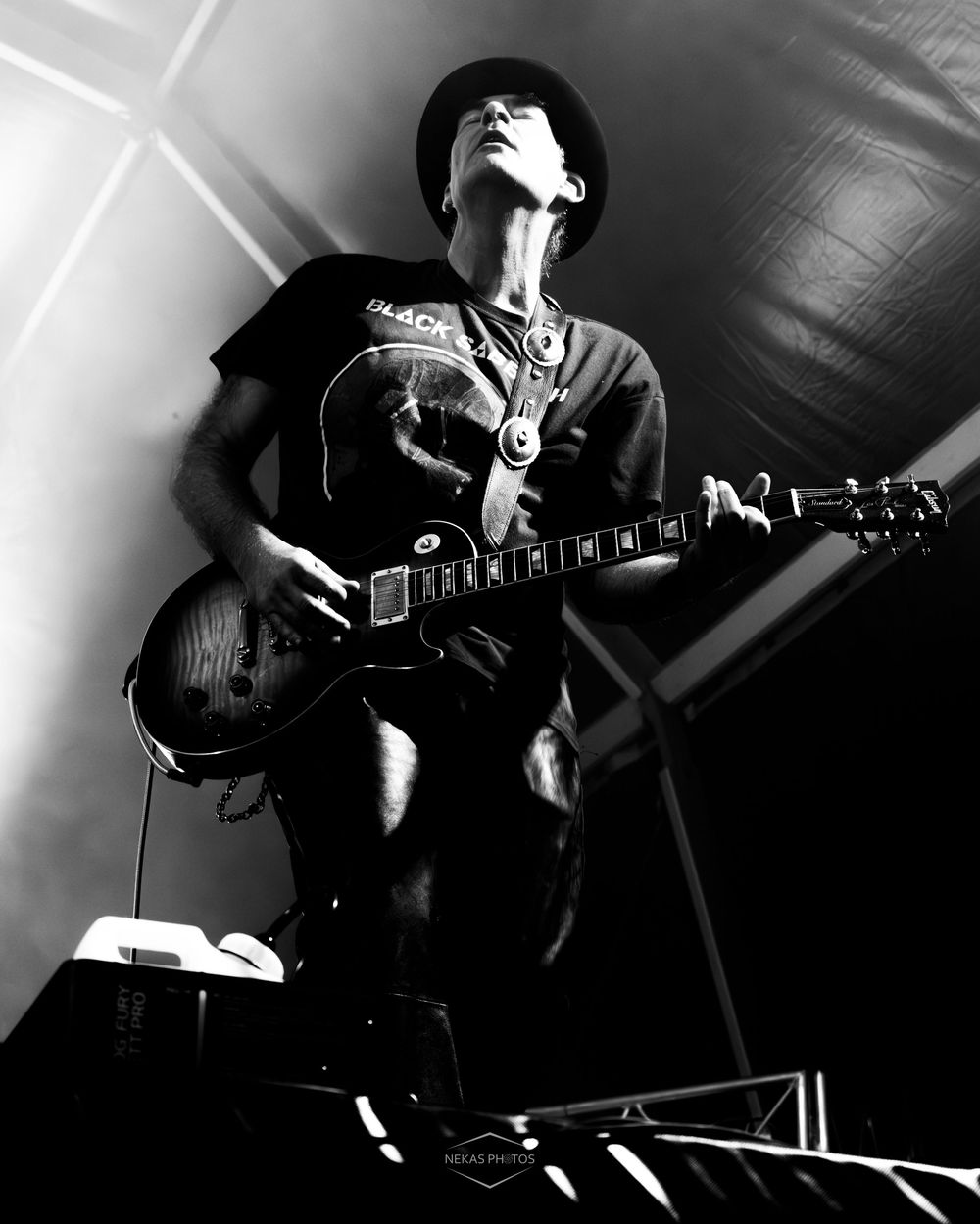 Black and white photo of a guitarist performing on stage, wearing a hat and t-shirt, playing an electric guitar under dramatic lighting.