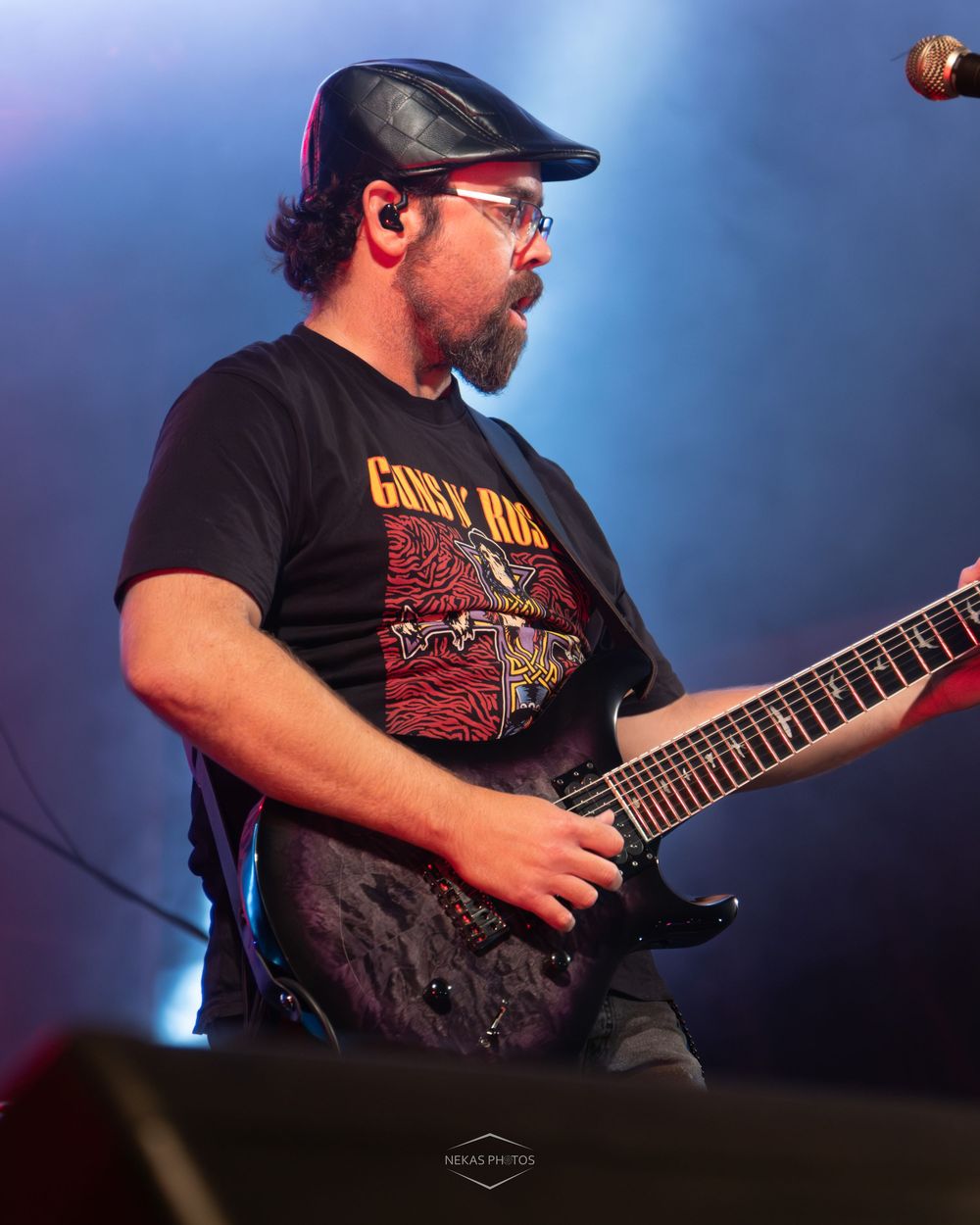 Bearded male guitarist in black band tee and flat cap performing electric guitar on stage under blue and purple lights.
