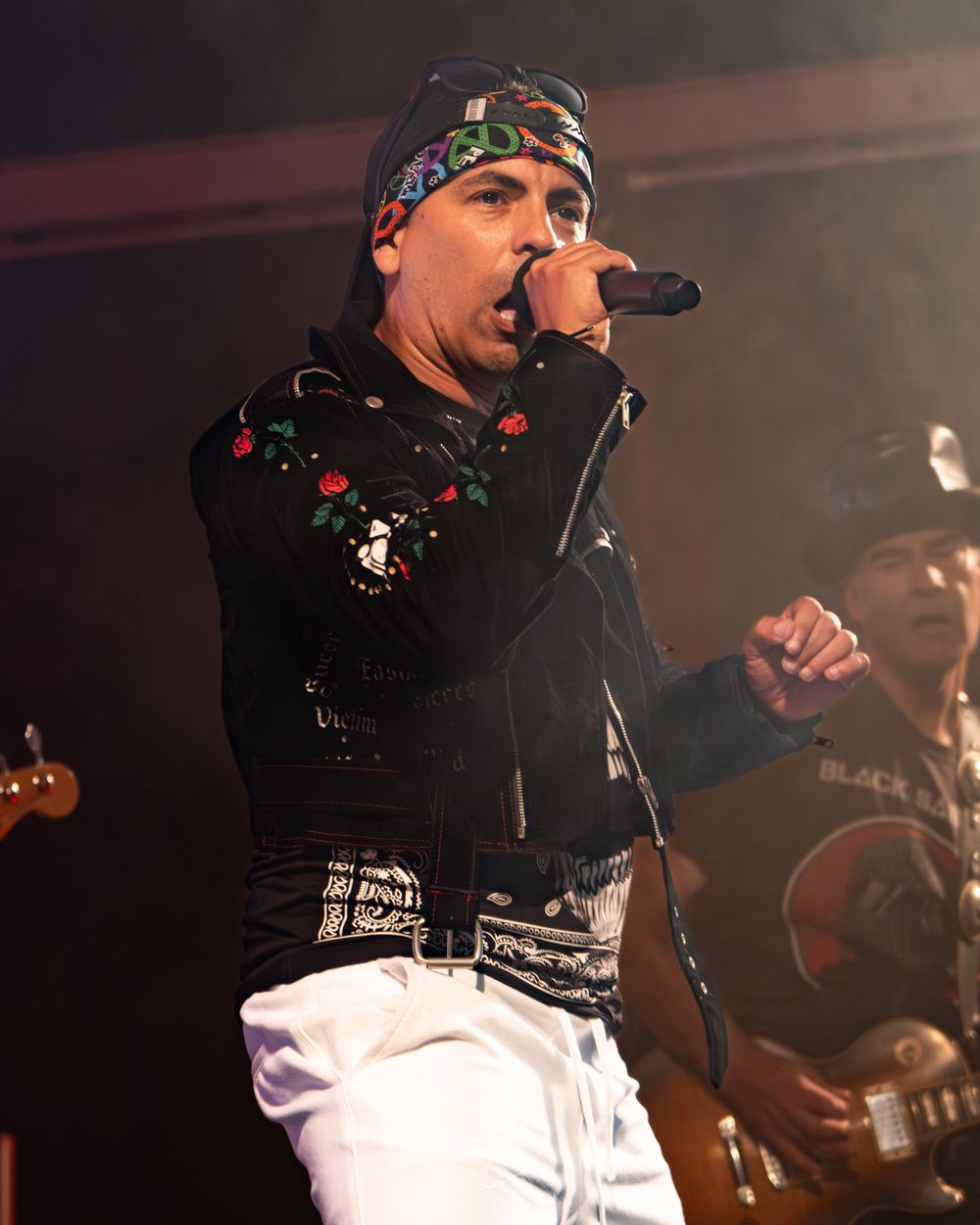 A rock singer performs on stage wearing a black embroidered jacket, bandana, and white pants while holding a microphone.