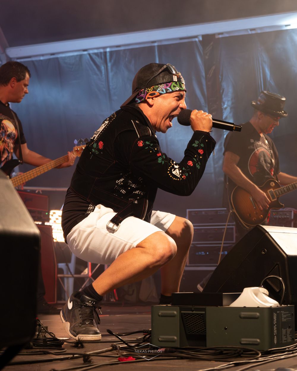 A rock band performs on stage, with an energetic male vocalist crouching and screaming into a microphone wearing a floral jacket.