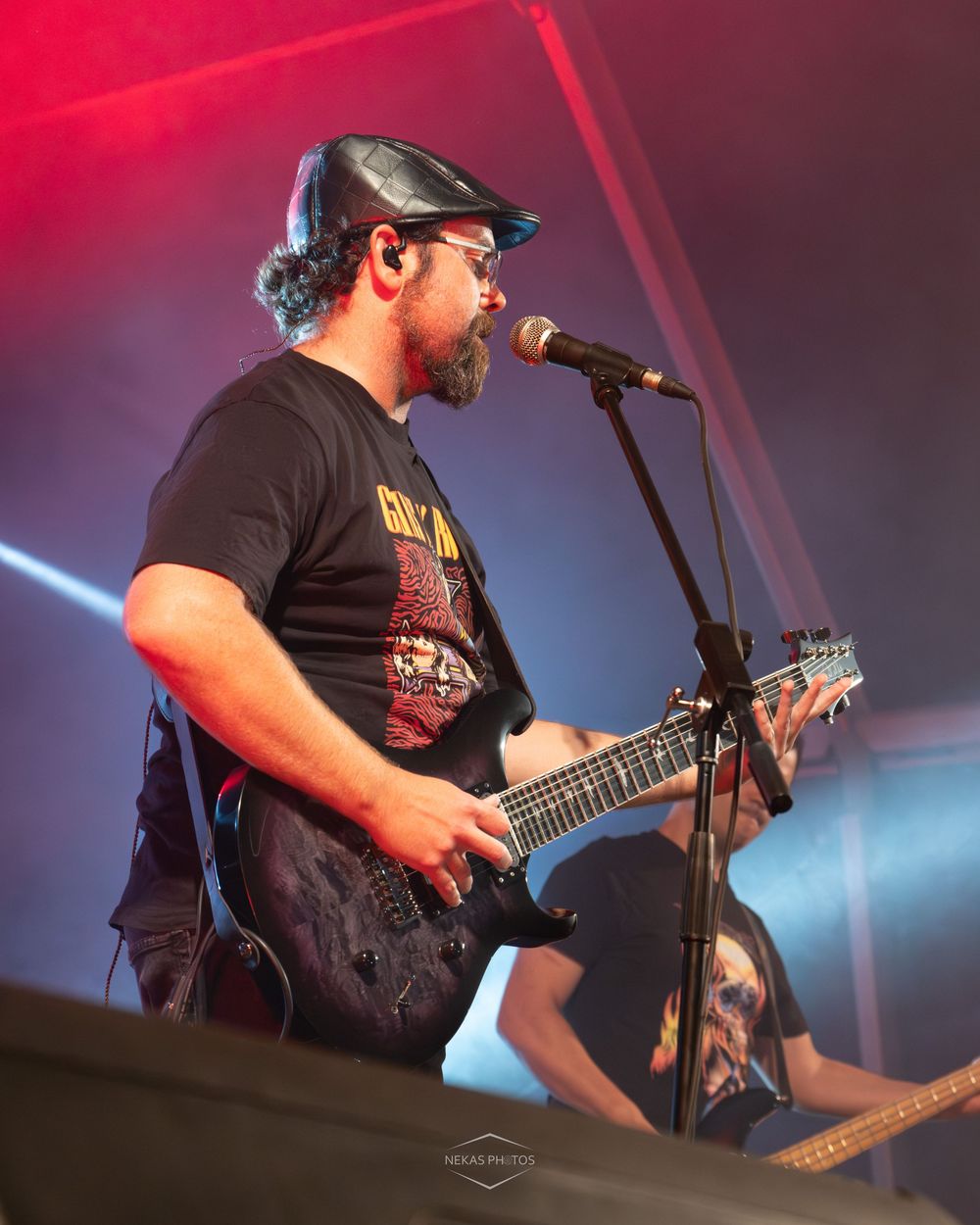 Bearded male guitarist performing on stage, wearing a black band tee and leather cap, playing electric guitar at a live concert.