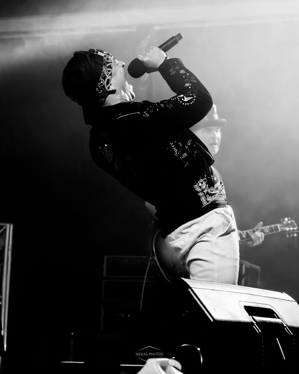 Black and white photo of a rock vocalist singing passionately on stage with a guitarist visible in the background.