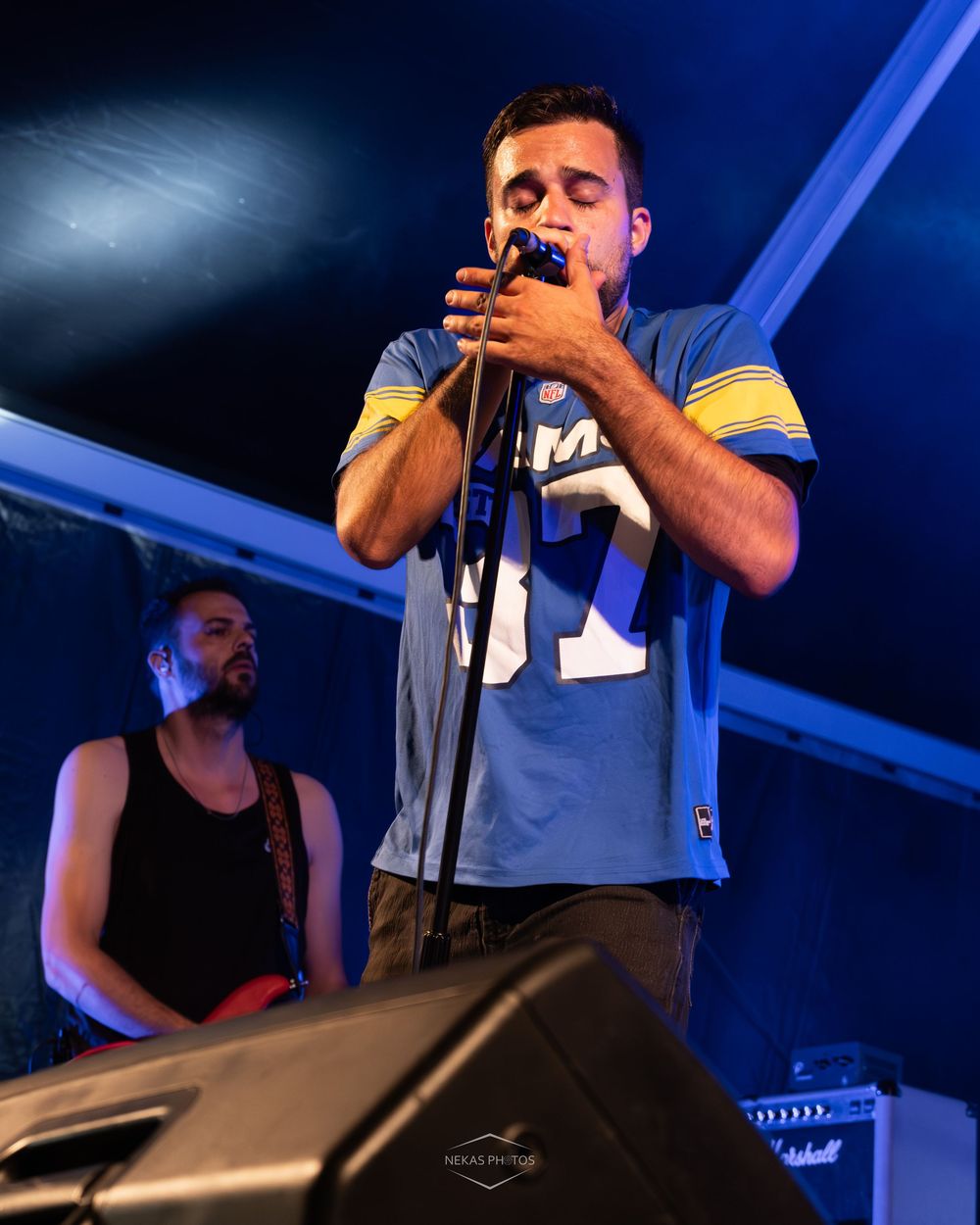 A male singer performs on stage holding a microphone, wearing a blue jersey, with a guitarist visible in the background.