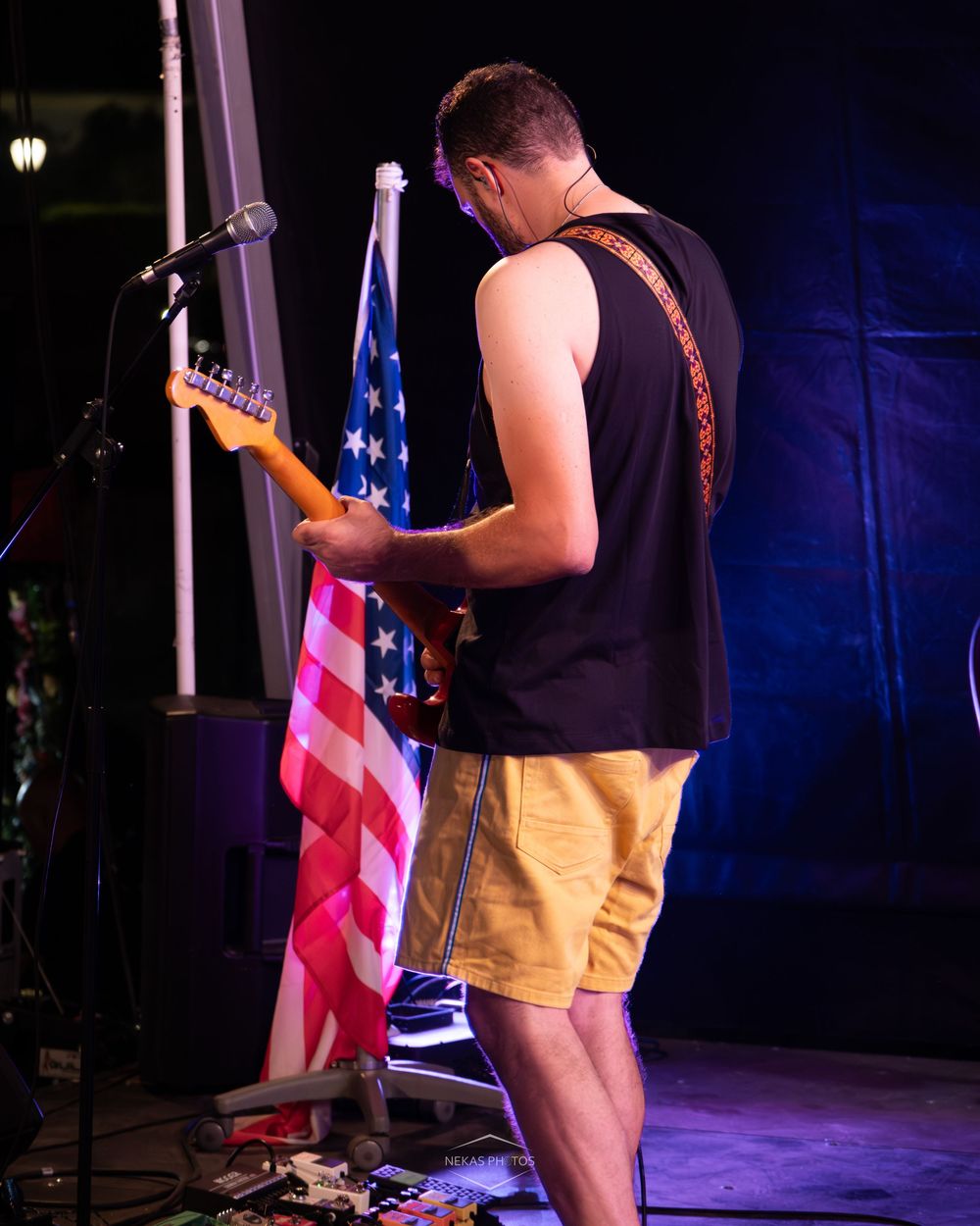 Guitarist performing on stage in black tank top and shorts, playing electric guitar in front of an American flag.