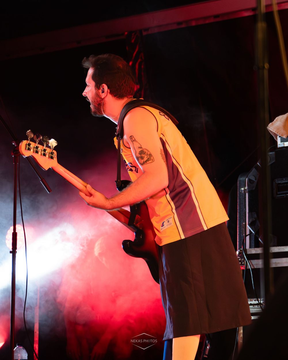 Male guitarist performing on stage with red fog lights, wearing a yellow jersey and playing a bass guitar at a live concert.