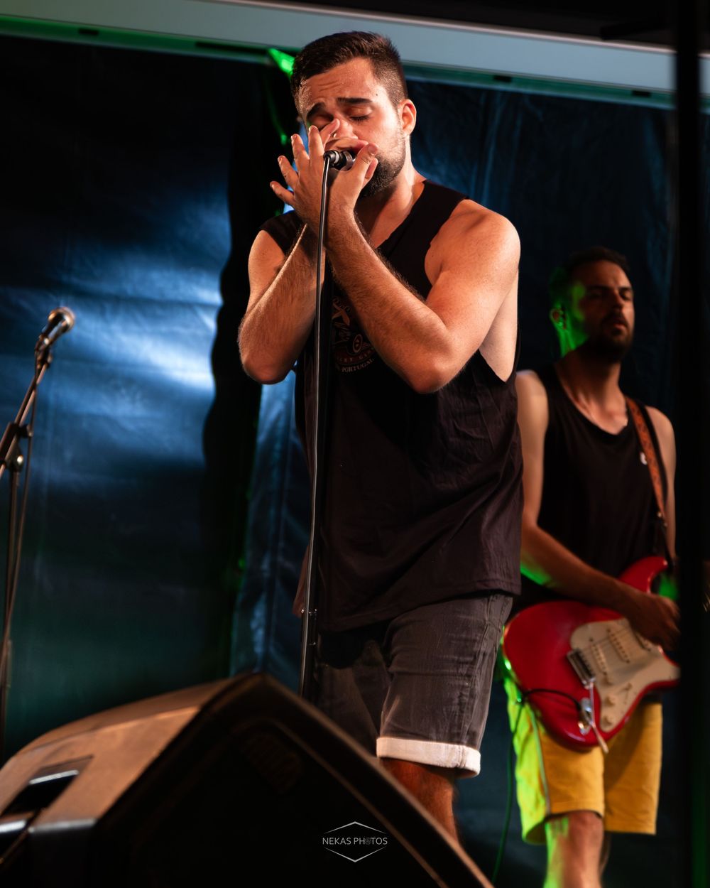 Male vocalist singing into microphone on stage with guitarist in background at live music concert.