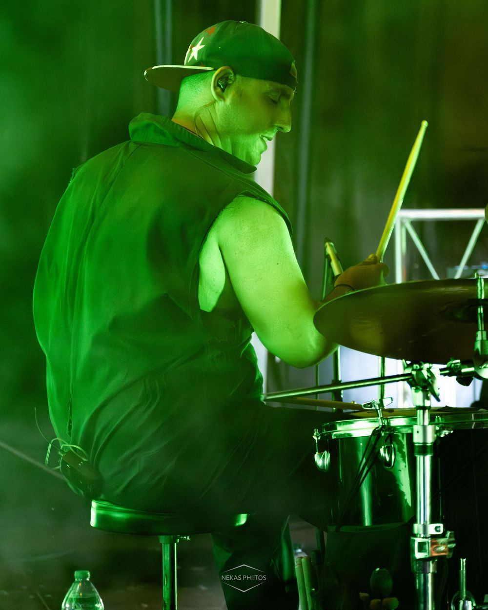 Drummer performing on stage under dramatic green lighting, playing cymbals with drumsticks while wearing a cap and vest.