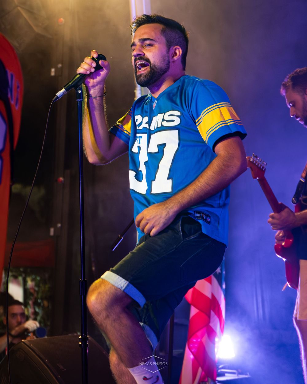 Man singing on stage wearing a blue Rams #37 jersey while performing live with a guitarist in the background.