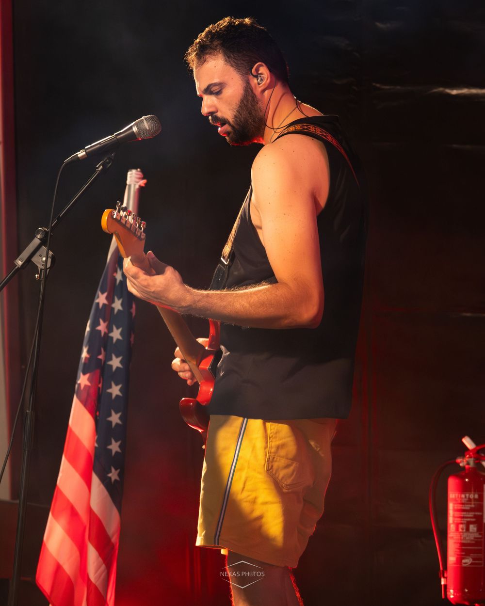 Male guitarist performing on stage with American flag backdrop, wearing yellow shorts and black sleeveless shirt, playing electric guitar.