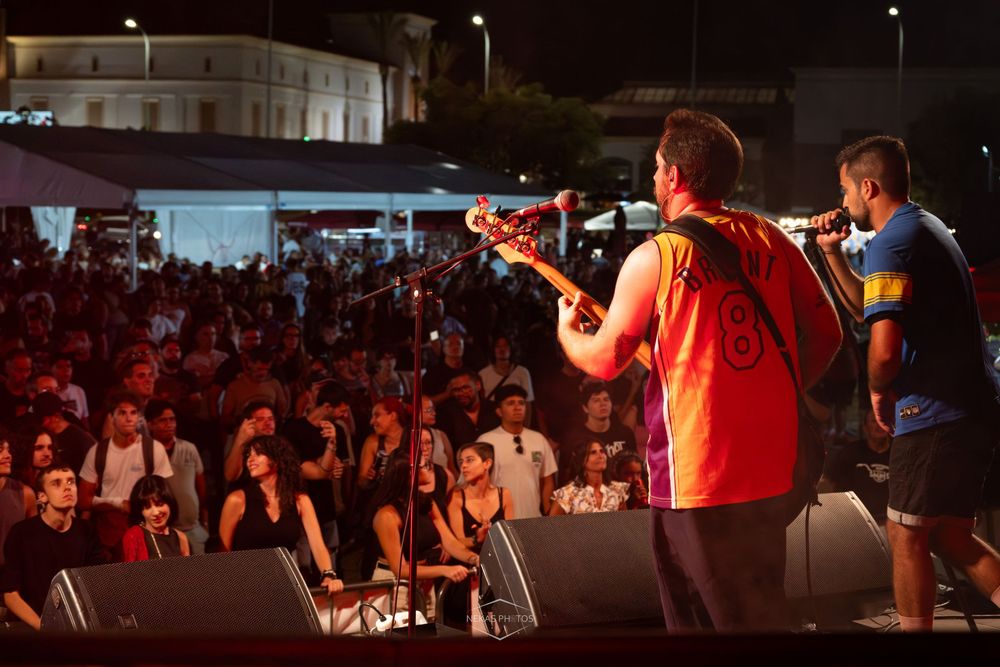 Band performing on outdoor stage at night with guitarist in red jersey playing to large crowd at summer concert.