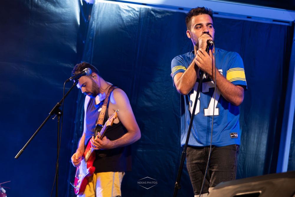 Two musicians performing on stage, one playing electric guitar and one singing into a microphone at a live event.