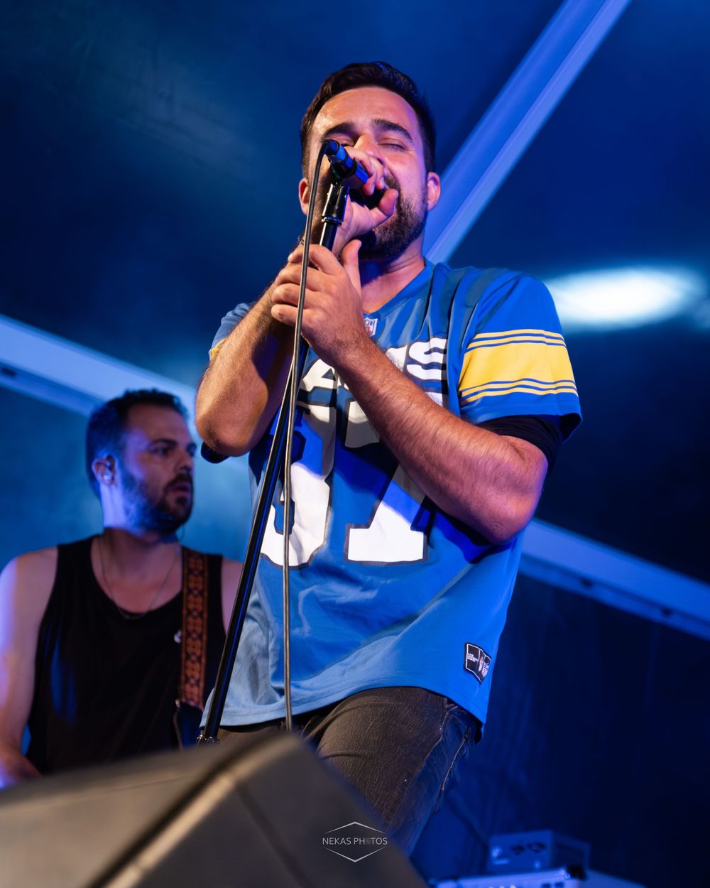 Male singer in blue football jersey performs on stage, gripping microphone stand with eyes closed under blue stage lighting.