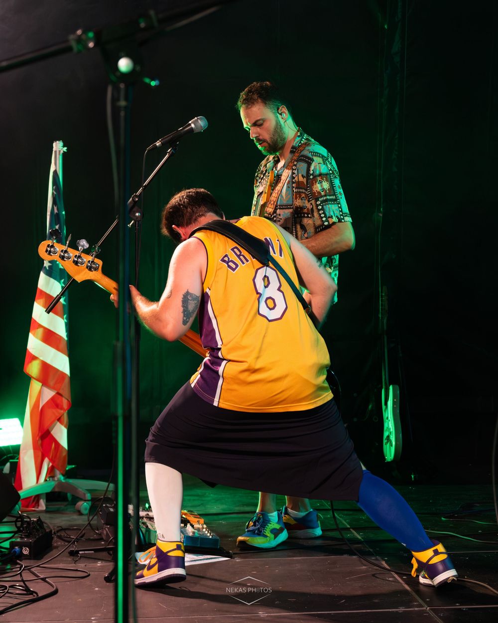 Two musicians perform energetically on stage, one wearing a Lakers jersey number 8, playing guitar under green stage lights.