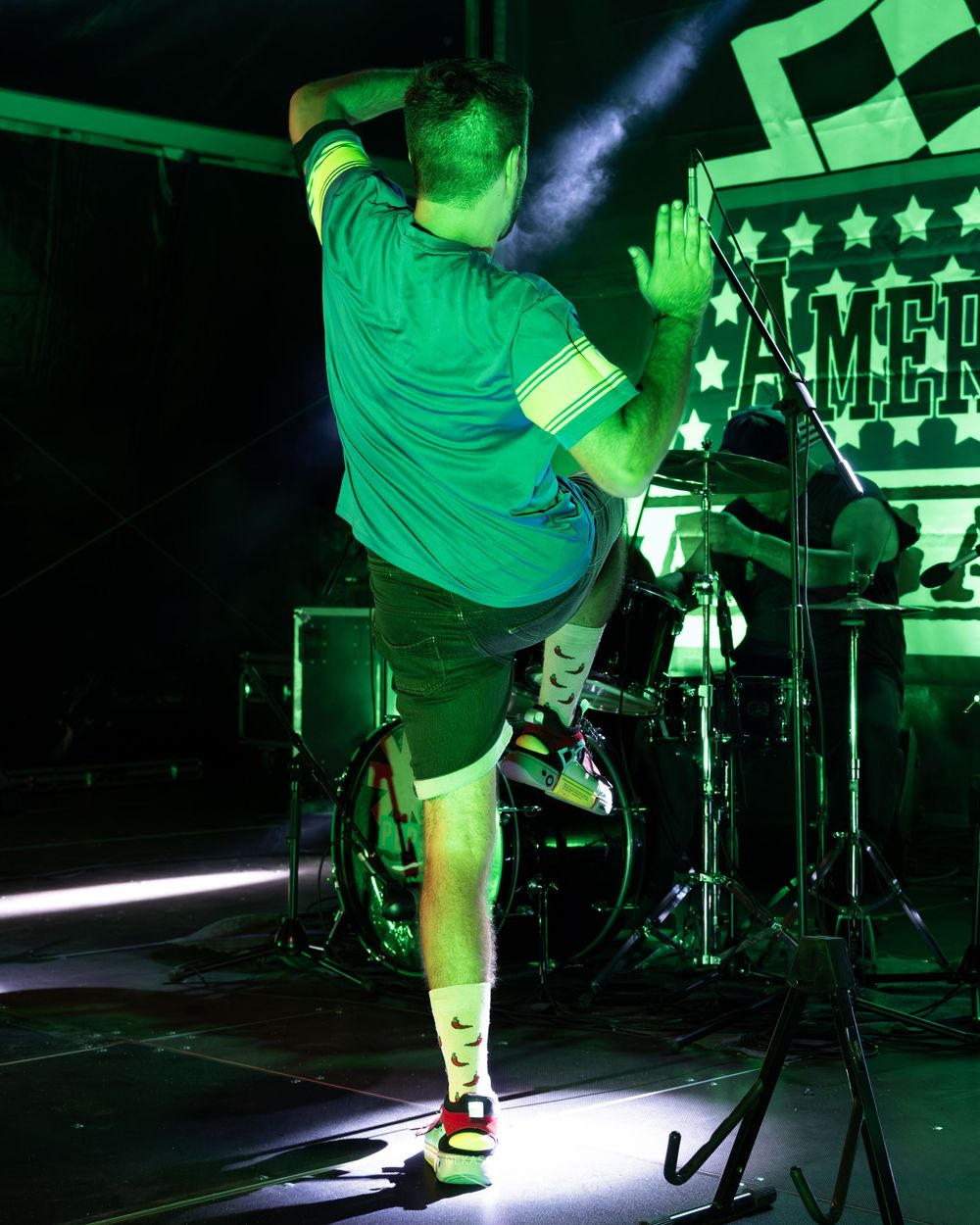 Performer in green shirt on stage under neon lights with drums and American-themed backdrop.
