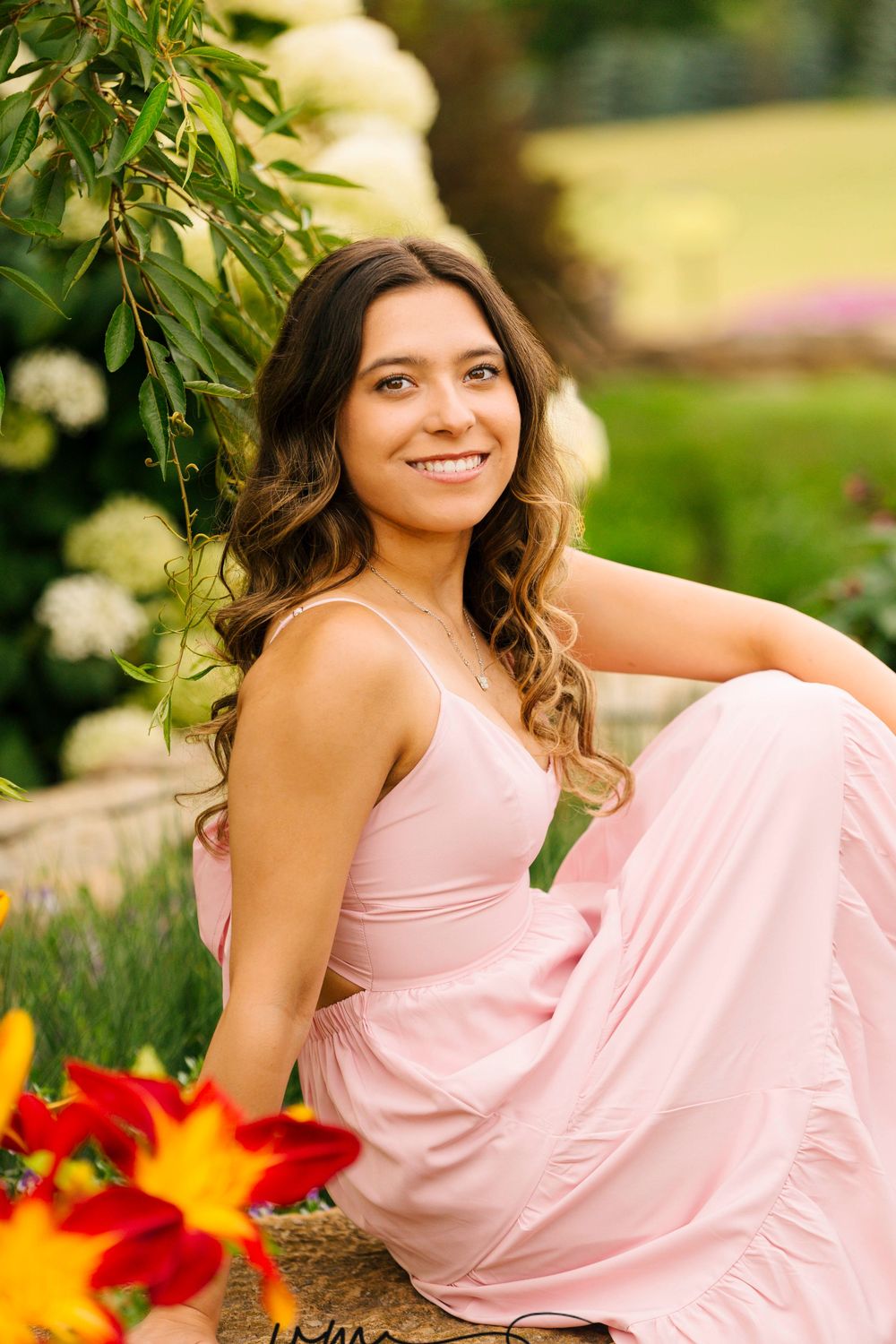 Young woman in pink maxi dress smiling outdoors among lush greenery and colorful flowers on a sunny day.