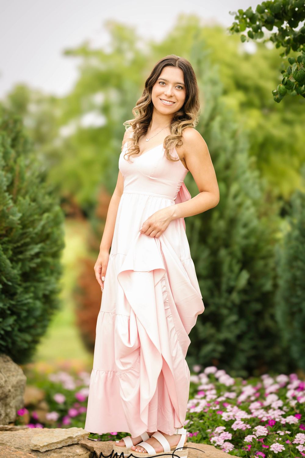 Young woman in a pink maxi dress smiling outdoors among green trees and colorful flowers.
