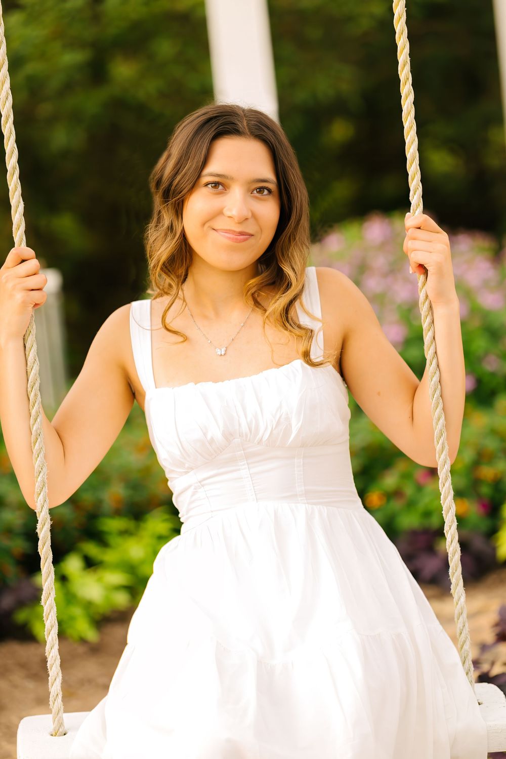 Young woman in white dress sitting on rope swing outdoors, smiling softly with garden flowers in background.
