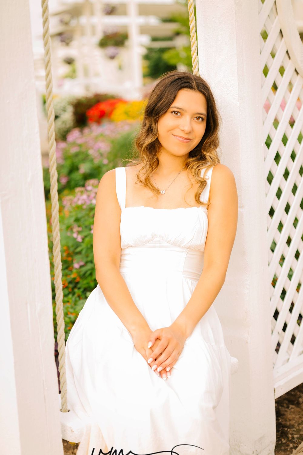 Young woman in white sundress sitting on a garden swing surrounded by colorful flowers and white lattice.