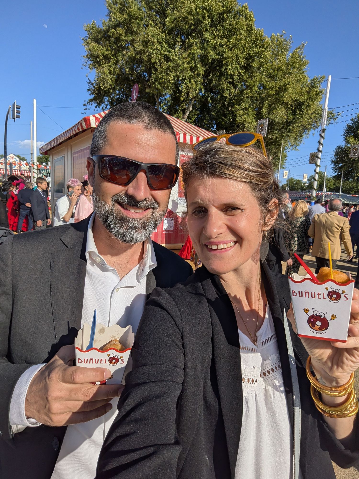 Couple smiling and holding buñuelos snacks at a festive outdoor fair with red and white striped booths.
